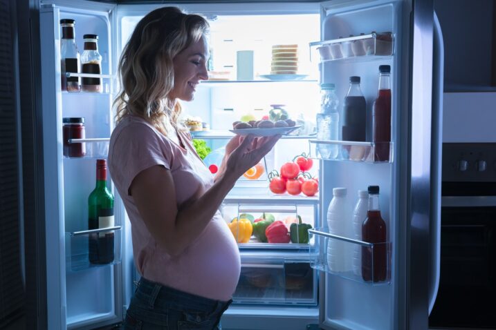 nosečnica stoji pred odprtim hladilnikom s krožnikom v rokah