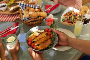 Pogled z visokega zornega kota na roke moškega, ki podaja krožnik s kuhanim piščancem in sedi za mizo med družinskim obrokom zunaj na terasi na soncu