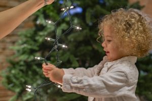 malček stoji ob božičnem drevesu in v rokah drži novoletne lučke