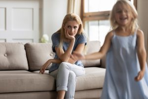 utrujena mama sedi na kavču, pred njo razigrana deklica