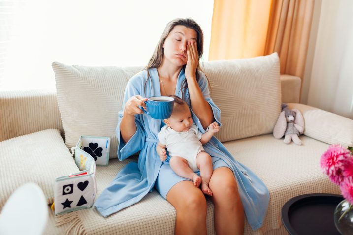 utrujena ženska sedi na kavču, pije kavo, v naročju ima dojenčka