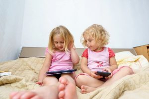 deklici sedita skupaj na postelji s tablico in telefonom v roki